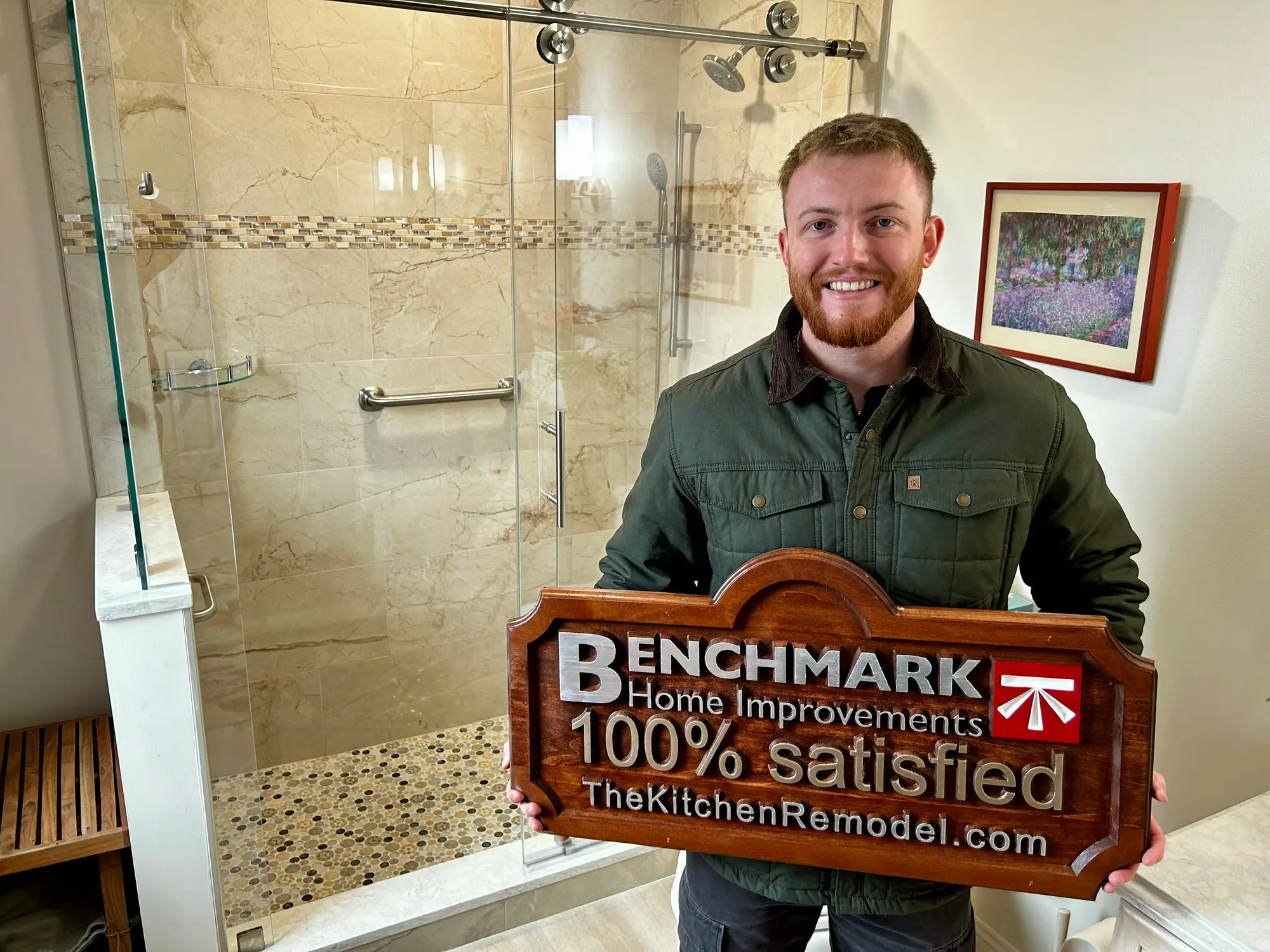 A wooden Benchmark “100% Satisfied” sign in a finished bathroom with a marble walk-in shower