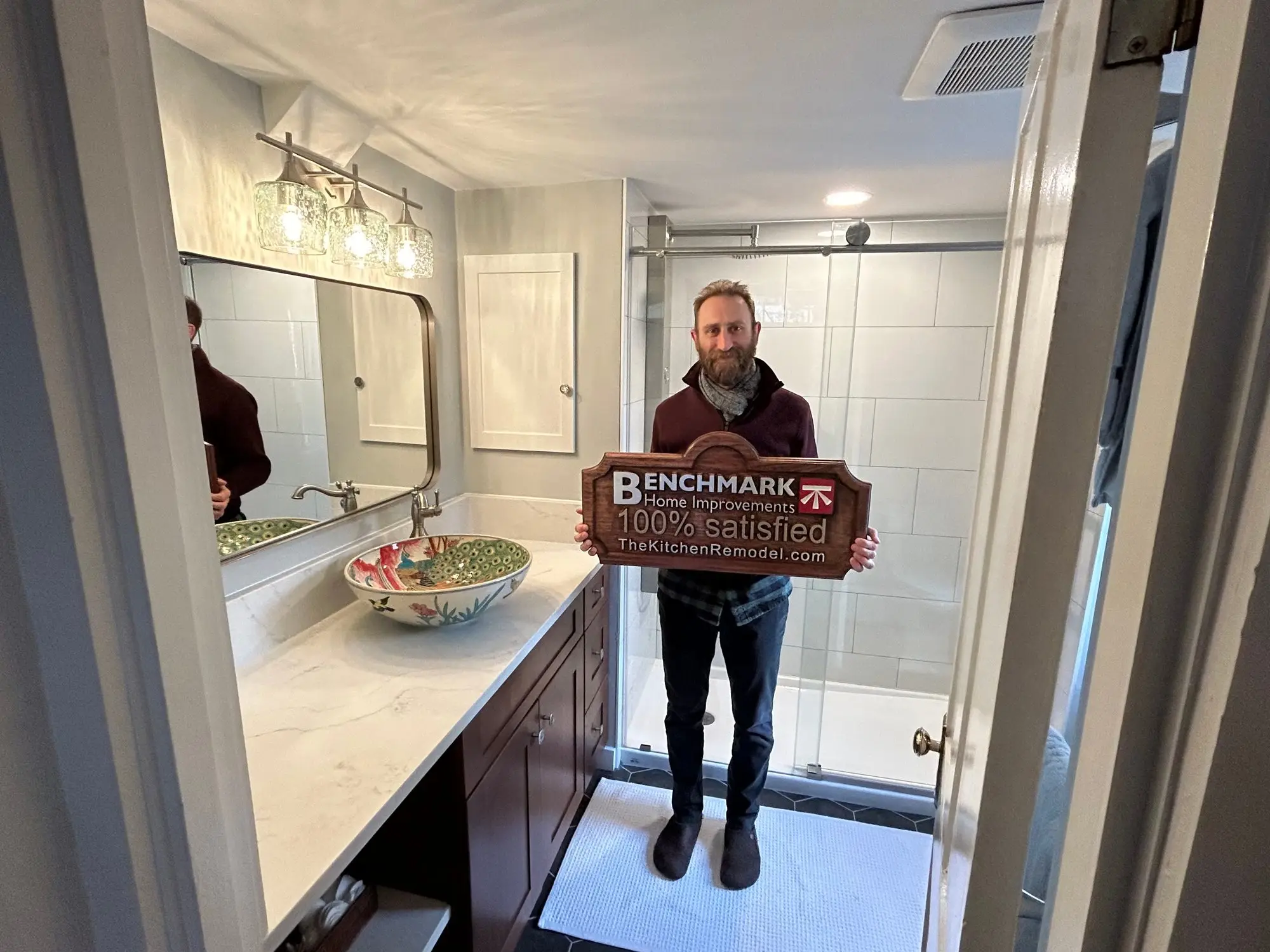 A homeowner holding a wooden Benchmark “100% Satisfied” sign in a finished bathroom with a marble vanity and glass shower