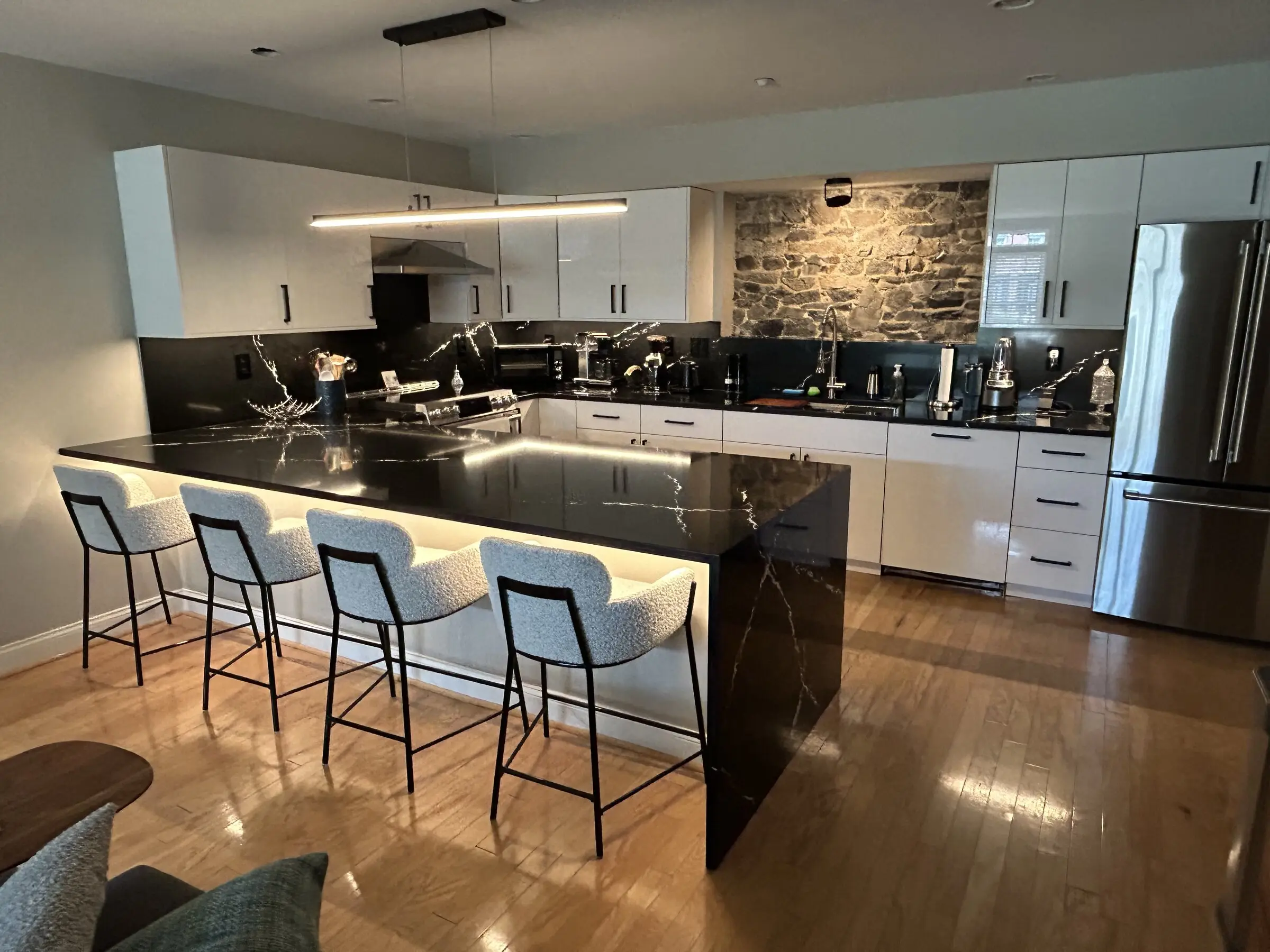 Modern kitchen with a black quartz waterfall island, white high-gloss cabinets, and a stone accent wall, remodeled by Benchmark on the New Hampshire Seacoast