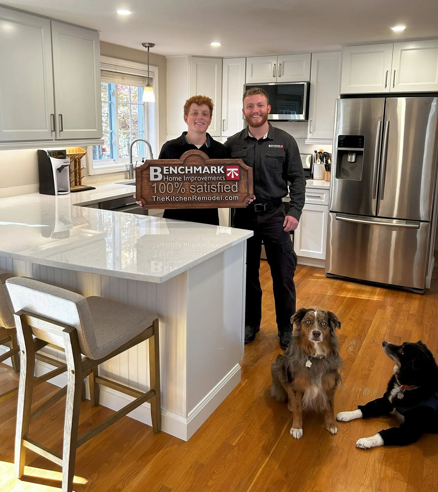 Two people holding a wooden Benchmark “100% Satisfied” sign in a finished white kitchen, with two dogs on the hardwood floor
