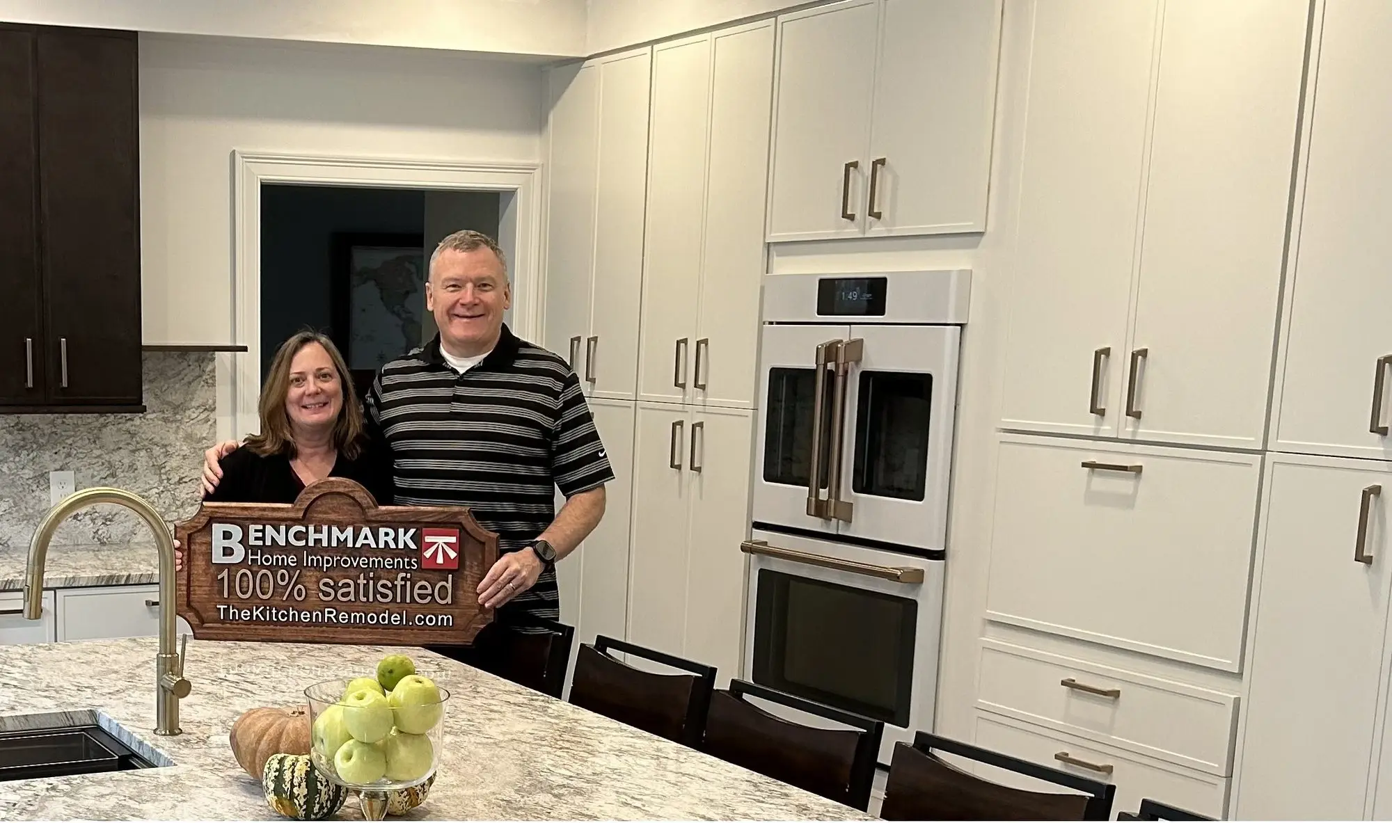 A homeowner couple holding a wooden Benchmark “100% Satisfied” sign in their finished white kitchen with granite counters