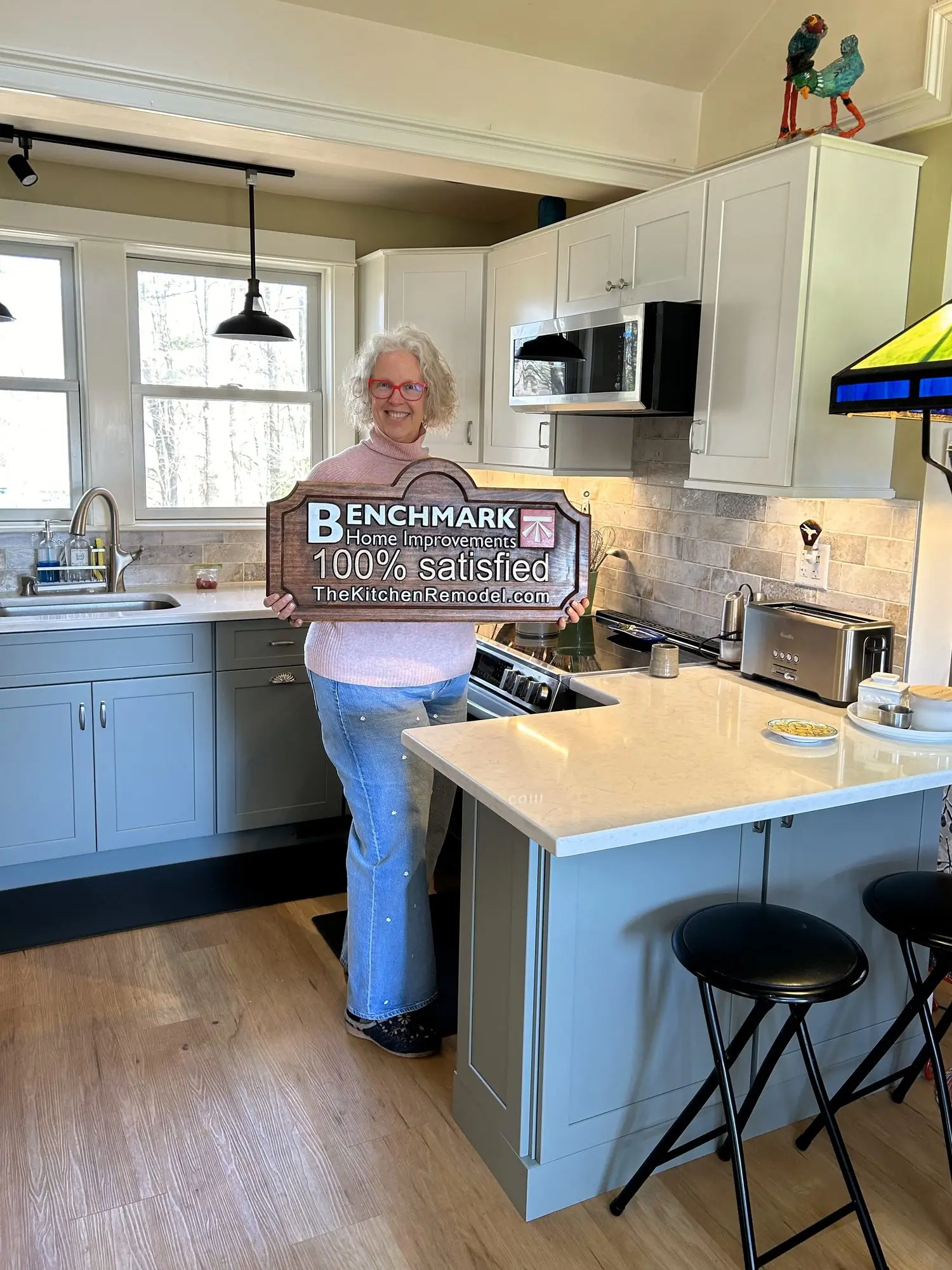 A homeowner holding a wooden Benchmark “100% Satisfied” sign in her finished sage-green kitchen