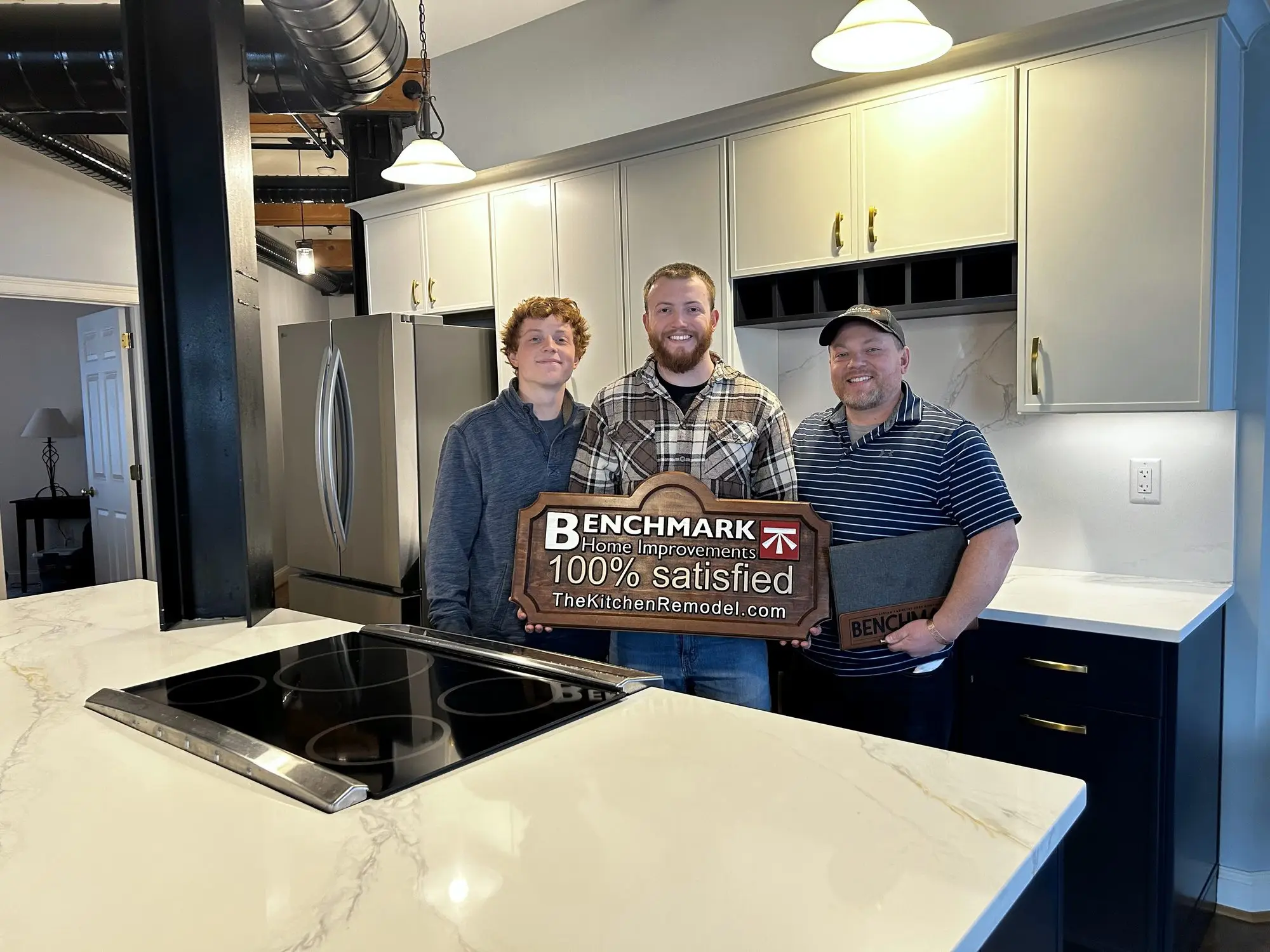 Three people holding a wooden Benchmark “100% Satisfied” sign in a finished kitchen with a white marble island and navy cabinets